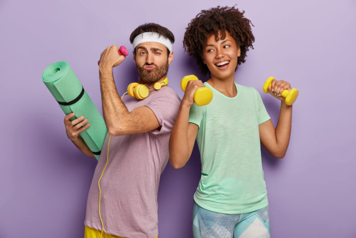 Glad multiethnic husband and wife attend sport center, exercise with dumbbells, hold fitness mat, stand back to each other, have funny happy looks, wear t shirts, isolated on purple background