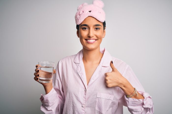 Young beautiful woman wearing pajama and sleep mask drinking glass of water happy with big smile doing ok sign, thumb up with fingers, excellent sign